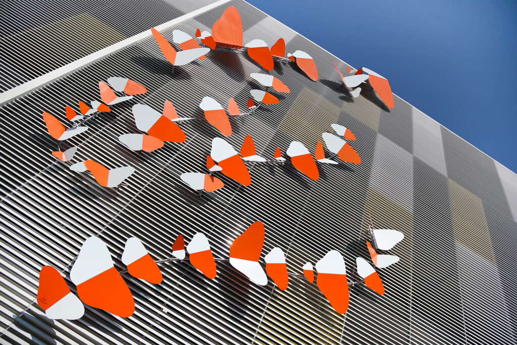 Moniosainen veistos, jossa erikokoisten, kaksiväristen perhosten runsaslukuinen parvi pyrähtää ylöspäin pitkin parkkihallin seinää.