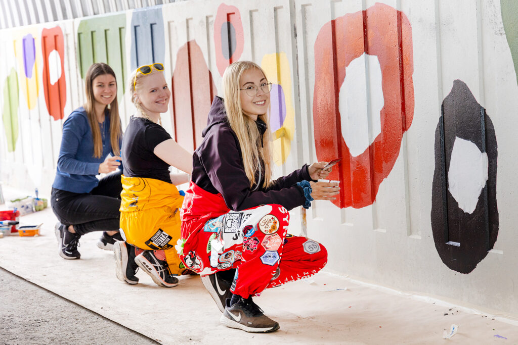 Kolme haalaripukuista opiskelijaa maalaa alikulkutunnelin seinään graafisia kuvioita.