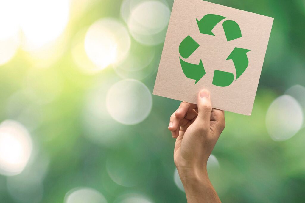 Käsi pitää ylhäällä pahvinpalaa, jossa on kierrätys-merkki. Taustalla vihreää luontoa.