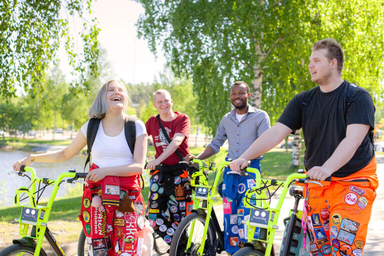 Neljä iloista, haalariasuista opiskelijaa on liikkeellä Vilkku-fillareilla.