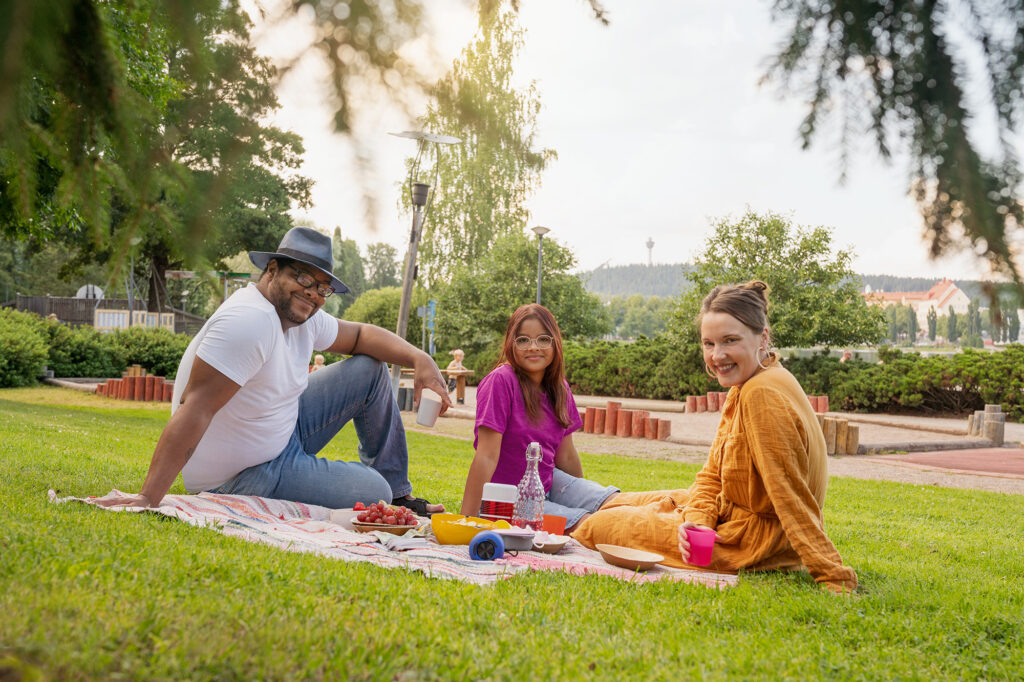Kolme iloista ihmistä piknikillä. Taustalla Puijon torni.