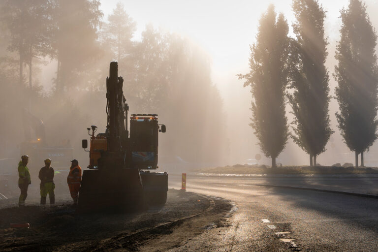 Kolme rakennusmiestä ja kaivinkone aamu-usvassa.