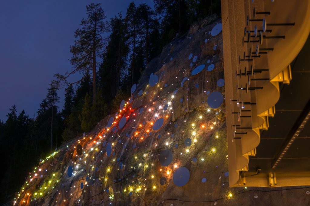 Pulssi-taideteos värivaloissa ilta-aikaan. Teos koostuu lukuisista kallioon kiinnitetyistä ympyränmuotoisista teräslevyistä ja led-valoista.